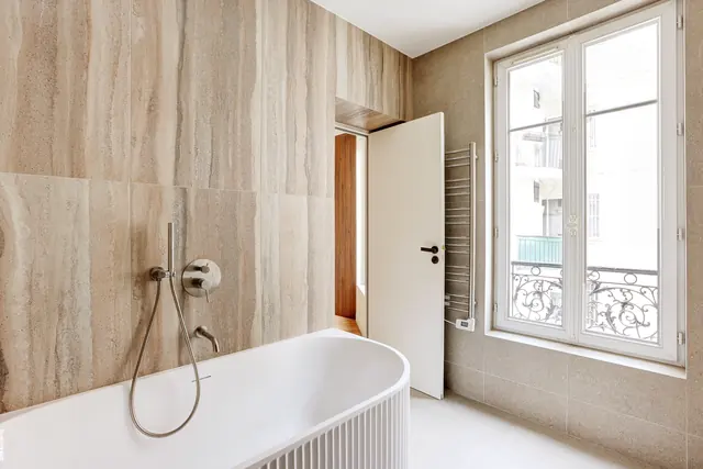 Salle de bains en travertin avec baignoire îlot et fenêtre sur rue parisienne